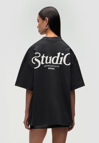 T-shirt noir surdimensionné avec un texte blanc au dos indiquant "Studio" et "Crafting Aesthetics". Tissu doux et coupe décontractée.