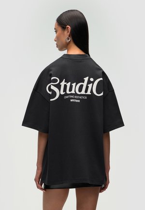 T-shirt nero oversize con testo bianco sulla schiena che recita "Studio" e "Crafting Aesthetics". Tessuto morbido e vestibilità rilassata.