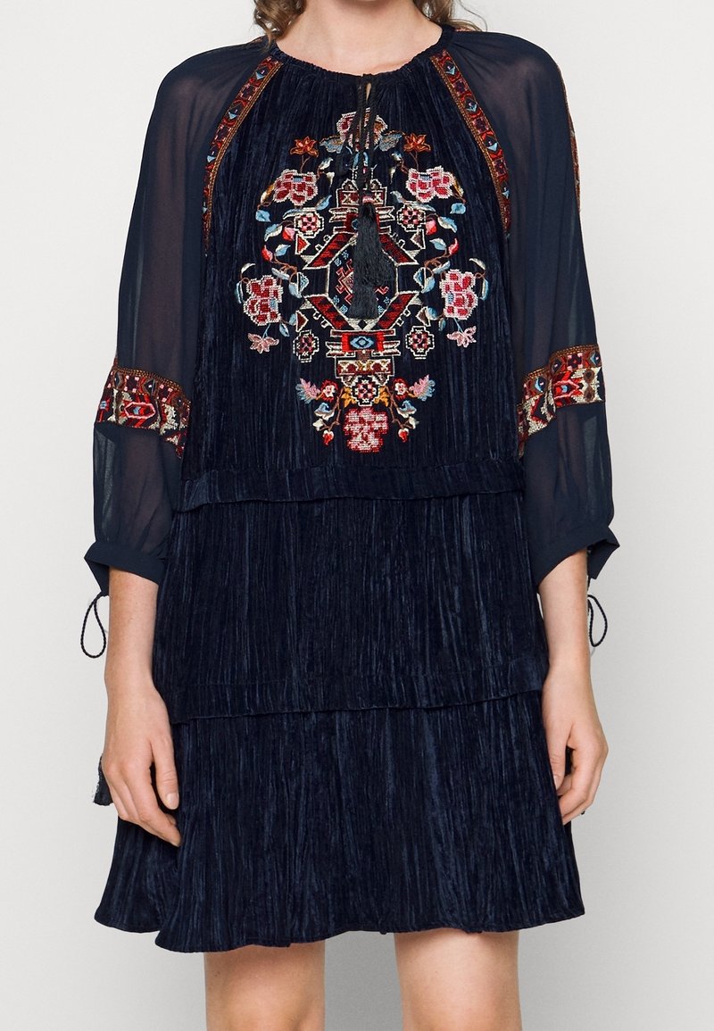 Robe bleu marine à volants avec un motif floral brodé sur le corsage, des manches semi-transparentes et un lien à pompon au niveau du cou. Tissu texturé.
