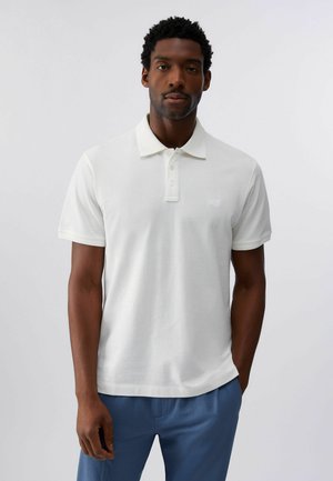 Uomo in piedi di fronte, indossa una polo bianca a maniche corte e pantaloni blu su uno sfondo chiaro e semplice.