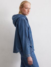 Blaue Jeansjacke mit Kapuze, lässiger Schnitt, auffällige Nähte, Brusttasche und strukturierte Oberfläche. Kombiniert mit passenden blauen Hosen.