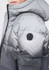 Chaqueta gris acolchada con capucha, que presenta un parche de logo negro en la manga, costuras horizontales y una textura de tela lisa y brillante.