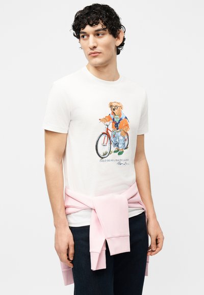 Νεαρός με κατσαρά μαλλιά φοράει λευκό μπλουζάκι με αρκουδάκι ντυμένο με casual ρούχα και ποδήλατο, ροζ φούτερ δεμένο γύρω από τη μέση, σκούρα παντελόνια.