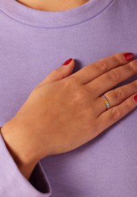Dunne gouden ring met meerkleurige hartvormige accenten, gedragen door een hand die op een lavendelkleurige stof rust. Gladde textuur en eenvoudig ontwerp.