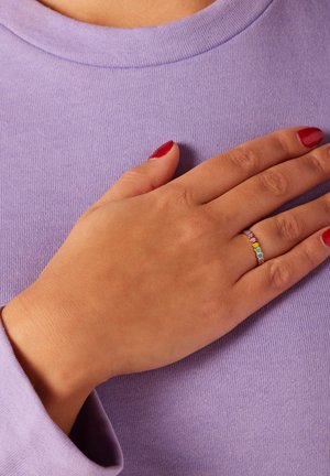Thin gold ring featuring multicolored heart-shaped accents, worn by a hand resting on a lavender fabric. Smooth texture and simple design.