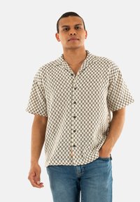 Camicia a maniche corte beige con motivo a quadretti neri, dotata di colletto a punta e chiusura con bottoni, abbinata a jeans in denim.