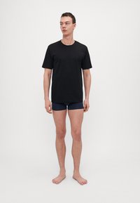 T-shirt in cotone nero con maniche corte e scollatura arrotondata, abbinato a biancheria intima blu navy, modello in piedi a piedi nudi contro uno sfondo semplice.