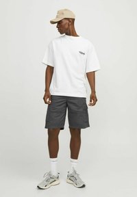 T-shirt blanc à manches courtes avec du texte à l'avant, assorti à un short cargo gris. Une casquette beige, des baskets grises et des chaussettes blanches complètent le look.