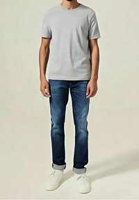 Homme portant un t-shirt gris clair à manches courtes, un jean bleu foncé retroussé et des baskets blanches, debout devant un fond uni.