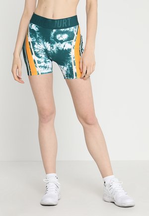Mujer con pantalones cortos de ciclismo tie-dye verdes y blancos con rayas naranjas, calcetines blancos y zapatillas deportivas blancas sobre un fondo liso.