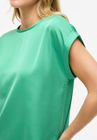 Blusa a maniche corte di un vivace verde, realizzata in tessuto morbido. Presenta un colletto rotondo e delle sottili aperture laterali per un maggiore comfort.