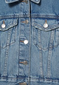 Veste en denim bleu clair vue de face avec des boutons-pression argentés, deux poches à rabat sur la poitrine et des détails de couture orange visibles.