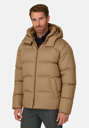 Homme portant une doudoune beige à capuche, une couche intérieure bleu marine et un pantalon gris, debout devant un fond clair uni.