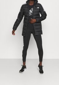 Veste matelassée noire avec capuche, rayures blanches sur les manches, associée à un t-shirt noir et des leggings noirs avec un détail logo blanc.