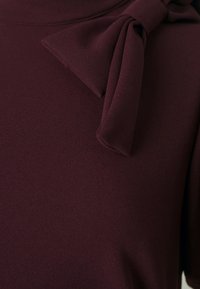 Burgundi felső, puha, texturált felülettel. Kiemelkedő masni részlet található a vállen, amely dimenziót ad a dizájnnak.