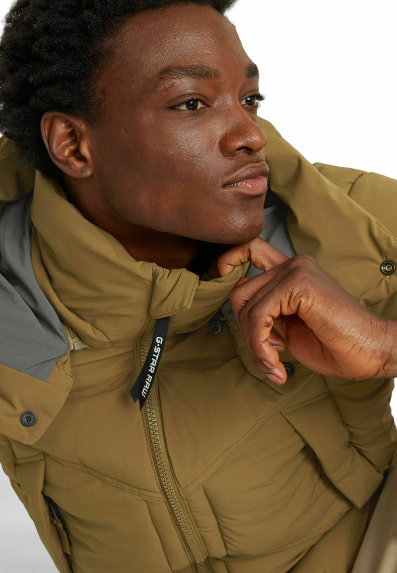 Kurtka puchowa w oliwkowozielonym kolorze z zamkiem błyskawicznym, wysokim kołnierzem i szarym akcentem na ramieniu. Posiada logo marki oraz detale z guzikami.