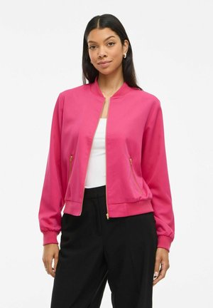 Woman wearing bright pink zip-up bomber jacket over white top and black pants, standing against white background.