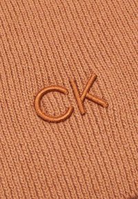 Kootud kangas sooja oranži tooni, millel on sooniline tekstuur. Esitleb pisut tumedamat tooni tikandiga "CK" logo.