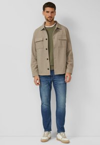 Mężczyzna w beżowej, zapinanej na guziki kurtce, oliwkowym swetrze, niebieskich dżinsach i białych tenisówkach stoi na jednolitym tle.