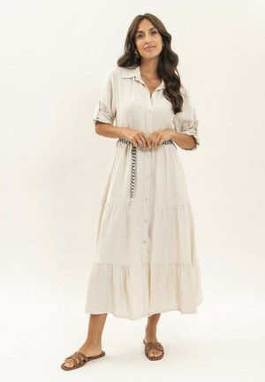 Femme portant une longue robe beige boutonnée avec des manches retroussées et une ceinture noire et blanche, debout avec des sandales plates marron.