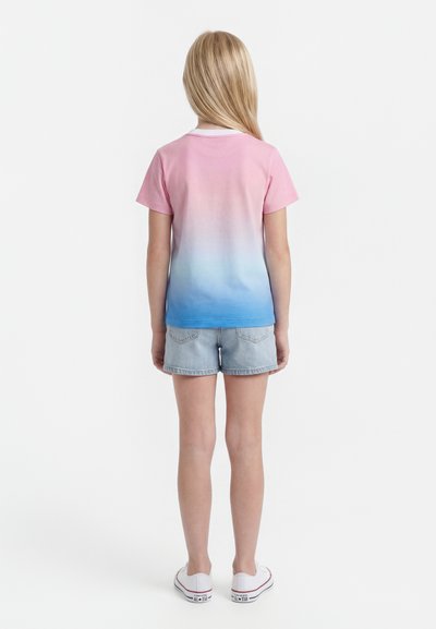 Παιδί με μακριά ξανθά μαλλιά που φοράει ροζ προς μπλε ombré μπλούζα, ανοιχτόχρωμα τζιν σορτς και άσπρα αθλητικά παπούτσια, στέκεται με την πλάτη προς τον θεατή.