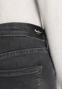 Slim-fit dunkelgraue Jeans mit glatter Textur, ausgestattet mit einem schwarzen Lederlabel, das "Pepe Jeans London" auf dem Bund geprägt ist.