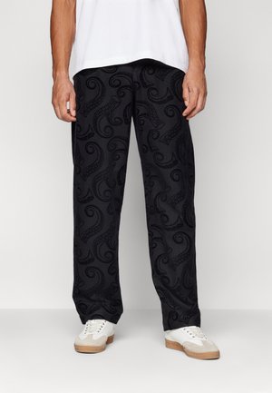 Mężczyzna ubrany w czarne spodnie z ciemnymi wzorami nawiązującymi do macek ośmiornicy oraz białe trampki, stojący na jasnym, jednolitym tle.