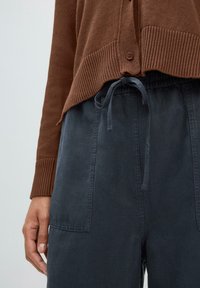 Cardigan en tricot marron avec des poignets côtelés et une fermeture à boutons, associé à un pantalon à cordon de serrage gris foncé avec des poches latérales.