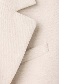 Gros plan sur le revers et la poche passepoilée d’un manteau en laine beige montrant un tissu doux et texturé.