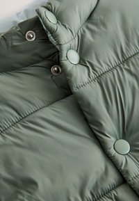 Gros plan sur un tissu de veste matelassée verte avec des lignes horizontales cousues et des boutons-pression, certains ouverts pour révéler les parties métalliques internes.