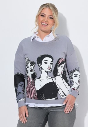 Smilende kvinde iført en grå sweatshirt med illustrerede portrætter af fem stilfulde forskellige kvinder på front og ærmer, kombineret med grå jeans.