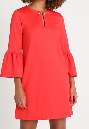 Vrouw draagt een koraalrode jurk met driekwart klokmouwen en een kleine sleutelgat-halslijn die bevestigd is met twee parelknopen.