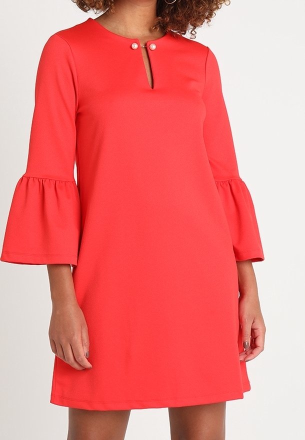 Femme portant une robe rouge corail avec des manches cloches trois quarts et un petit décolleté en forme de trou de serrure fermé par deux boutons en perle.