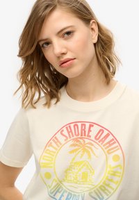 Camiseta de algodón color crema con un estampado gráfico colorido que incluye una palmera, una casa y texto, cuello redondo y mangas cortas.