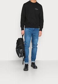 Sudadera negra, jeans azules y botas chelsea negras. Mochila negra de mano. Silueta casual con puños acanalados y cuello redondo.