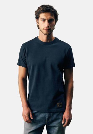 Mann, der nach vorne schaut, trägt ein schlichtes dunkelblaues T-Shirt mit kurzen Ärmeln und blaue Jeans vor einfachem hellem Hintergrund.