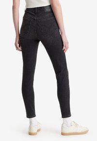 Personne portant un jean skinny taille haute noir, des baskets blanches et un haut blanc sans manches, vue de dos sur un fond blanc.