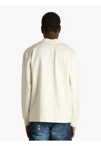 Felpa color crema con scollo rotondo e polsini a costine. Texture morbida, vestibilità comoda e design semplice sul retro. Abbinata a jeans blu strappati.