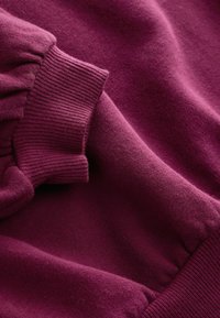 Primo piano di un ricco tessuto bordeaux con maniche texturizzate e polsini a costine, evidenziando la morbidezza e i dettagli del materiale.