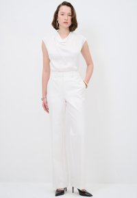 Top blanco sin mangas con un escote drapeado combinado con pantalones anchos blancos. La modelo lleva tacones negros de punta y joyería dorada sencilla.