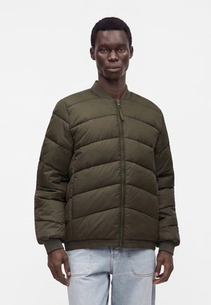 Homme portant une veste bomber matelassée vert olive foncé avec une fermeture éclair et un jean bleu clair, debout devant un fond blanc uni.