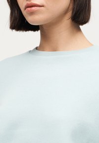 Femme aux cheveux courts et foncés portant un pull bleu clair à col rond, visible du menton jusqu'à la partie supérieure de la poitrine.