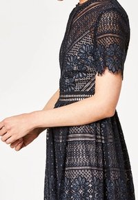 Navyblaues Spitzenkleid mit kurzen Ärmeln, das mit floralem Stickerei, strukturierten Mustern und einem taillierten Oberteil mit einem schwingenden Rockdesign versehen ist.