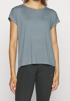 Femme portant un t-shirt gris ample à manches courtes et des leggings noirs moulants, debout avec les bras détendus le long du corps.