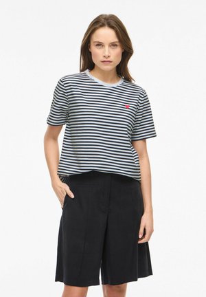 Kobieta ubrana w czarno-białą pasiastą koszulkę z krótkim rękawem, ozdobioną małym czerwonym sercem na piersi, w połączeniu z czarnymi szortami do kolan.