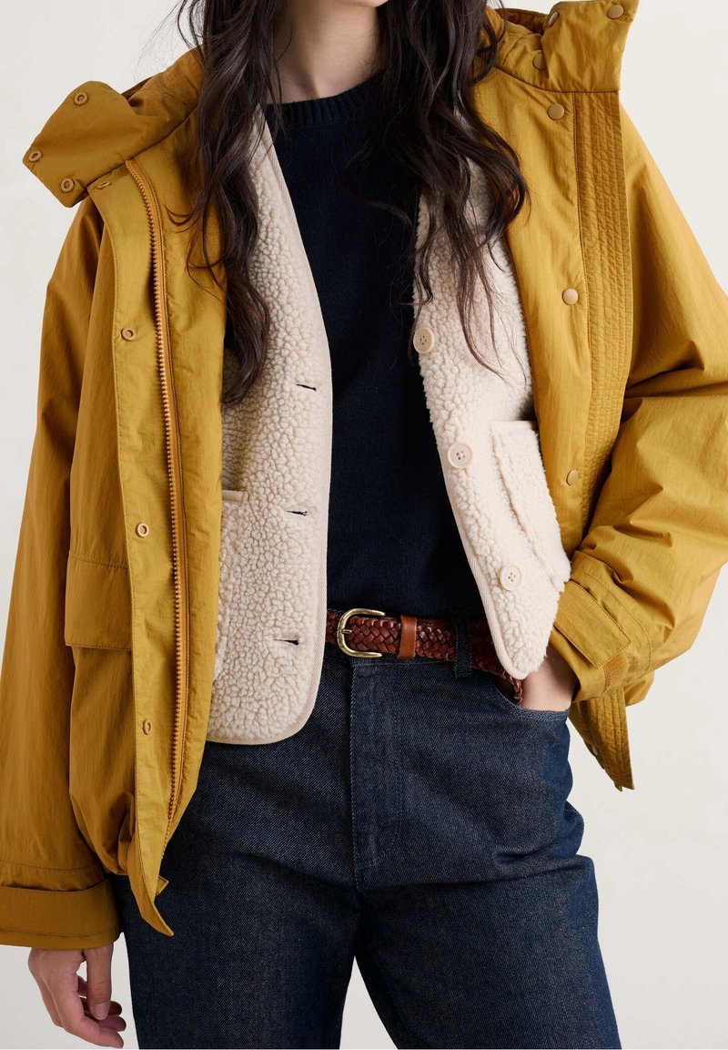 Femme portant une veste jaune moutarde sur un gilet sherpa crème, un haut noir, une ceinture tressée marron et un jean bleu foncé, main dans la poche.