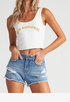 Vrouw draagt een wit cropped tanktop met de tekst "SUN GODDESS" en gescheurde blauwe denim shorts, handen in de zakken, tegen een lichte achtergrond.