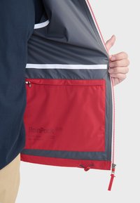 RainPack jack met een grijze binnenkant en een rode opbergzak, witte accentstrepen en een ritsmechanisme met een rode trekhaak.