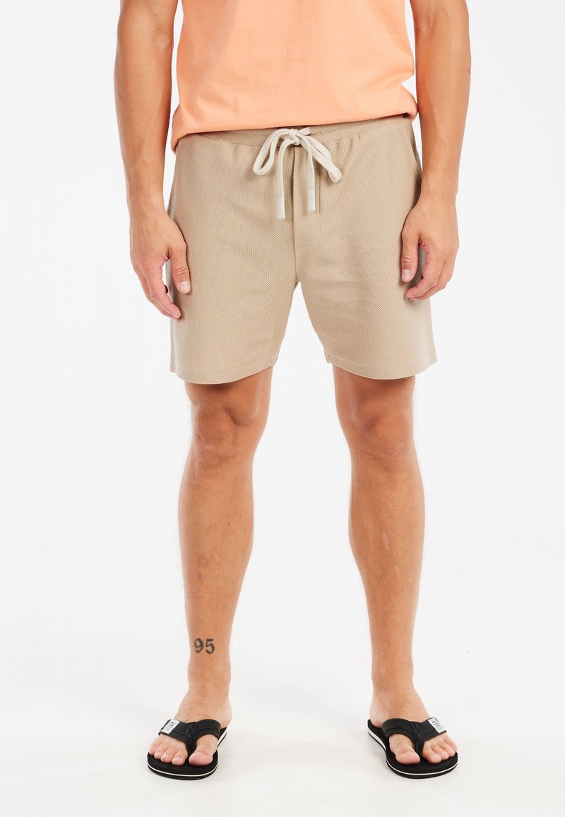 Beige strukturierte Shorts mit einem elastischen Bund und Kordelzug, kombiniert mit schwarzen Flip-Flops. Die Shorts haben eine Länge bis zur Mitte des Oberschenkels.