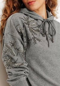 Sweat-shirt à capuche gris avec des ornements argentés détaillés sur les manches. Dispose d'une fermeture à cordon de serrage et d'une texture de tissu douce.